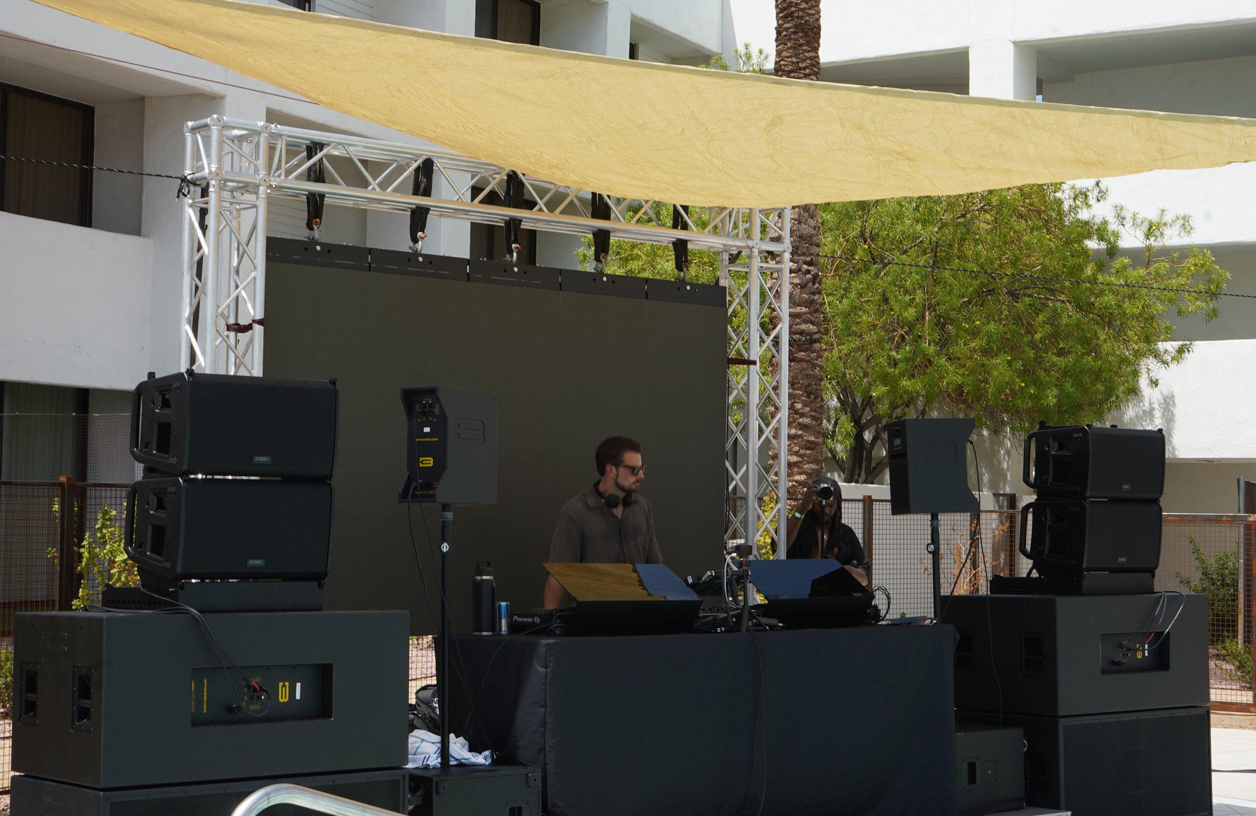The Arizona Sound System rig on a pool deck stage: line arrays on subwoofer stacks either side of a DJ booth under truss and a shade sail