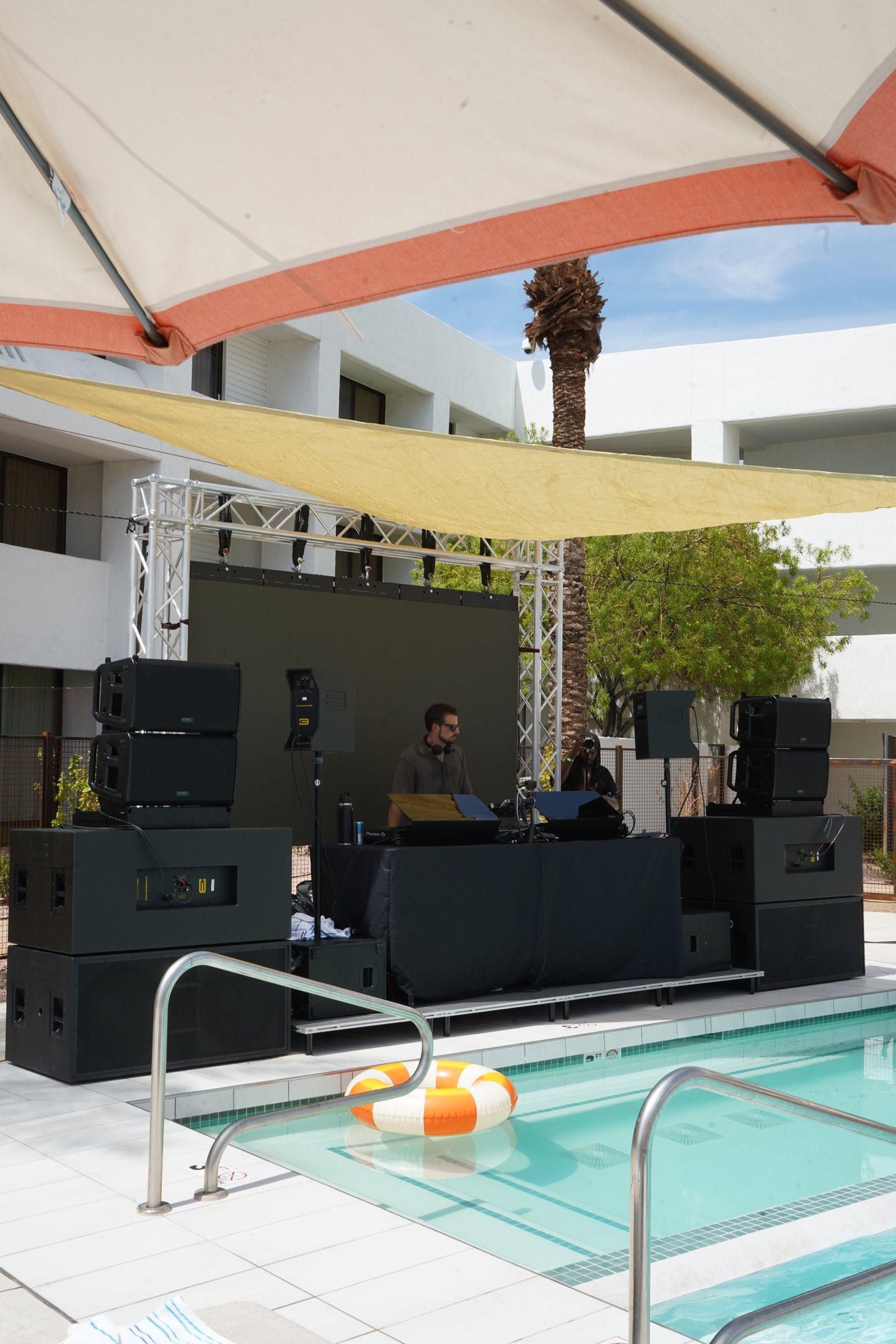 Line arrays stacked on subwoofers either side of a DJ booth under truss, beside a hotel pool